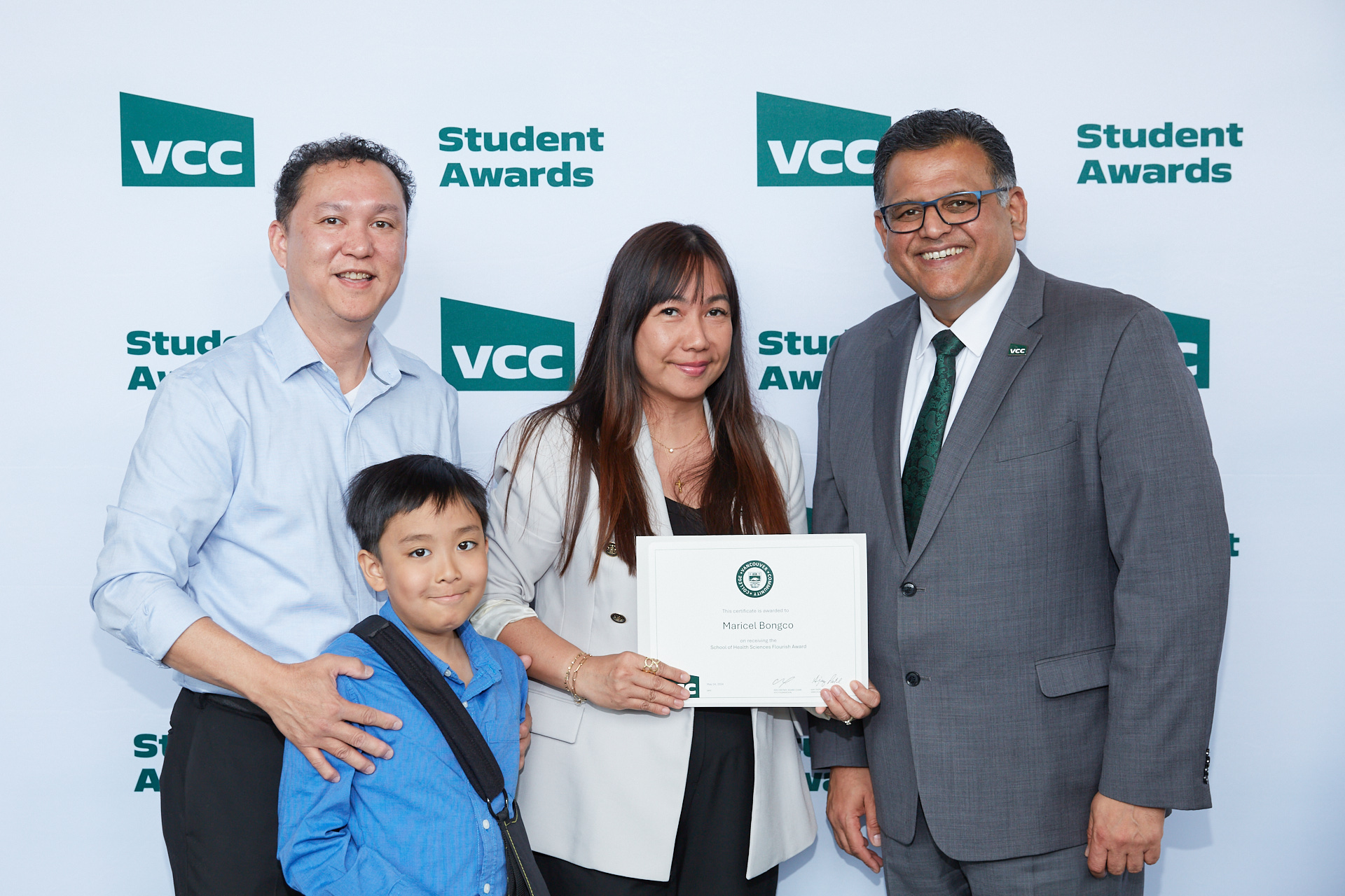 An award winner showing her certificate, with her family and VCC President by her side.