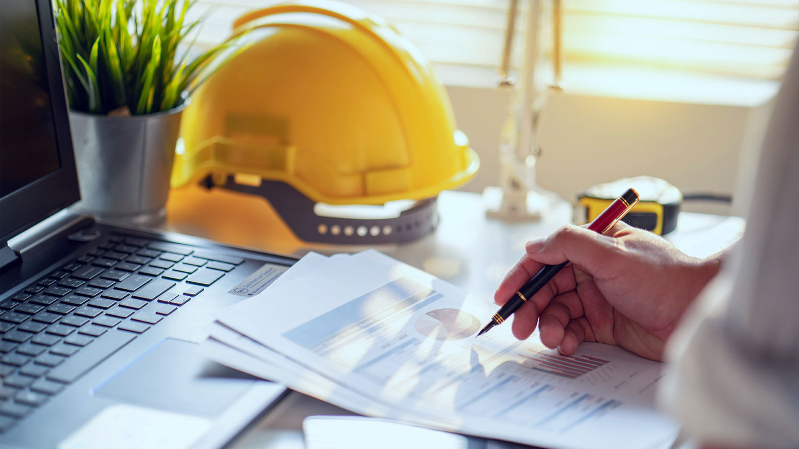 The construction person, with a hard hat nearby, uses a calculator and a laptop to manage the project