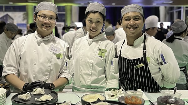 Three students chefs in white uniforms smiling behind a table with dishes.