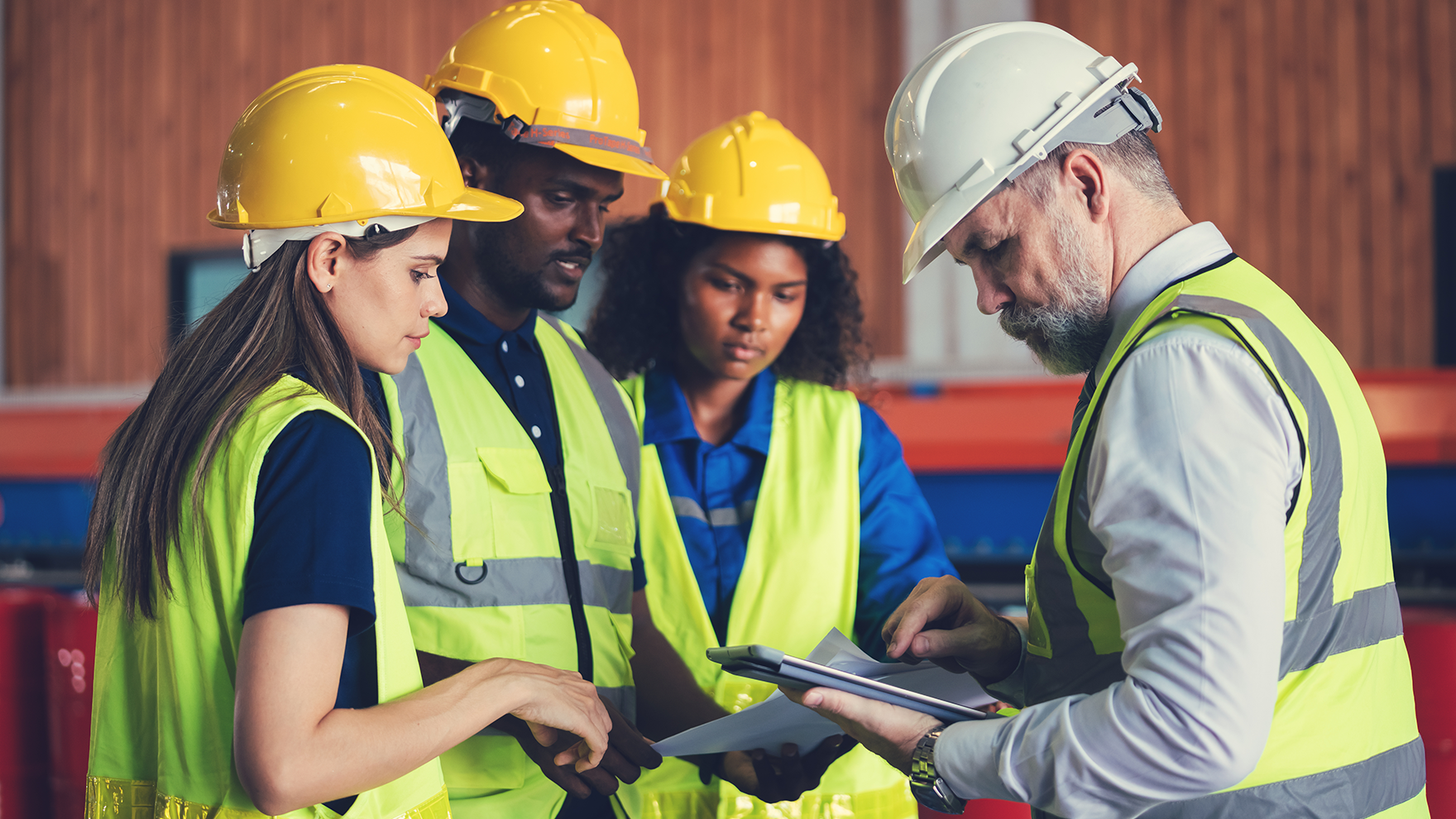 Four people in safety vests and hard hats review documents in a construction site.